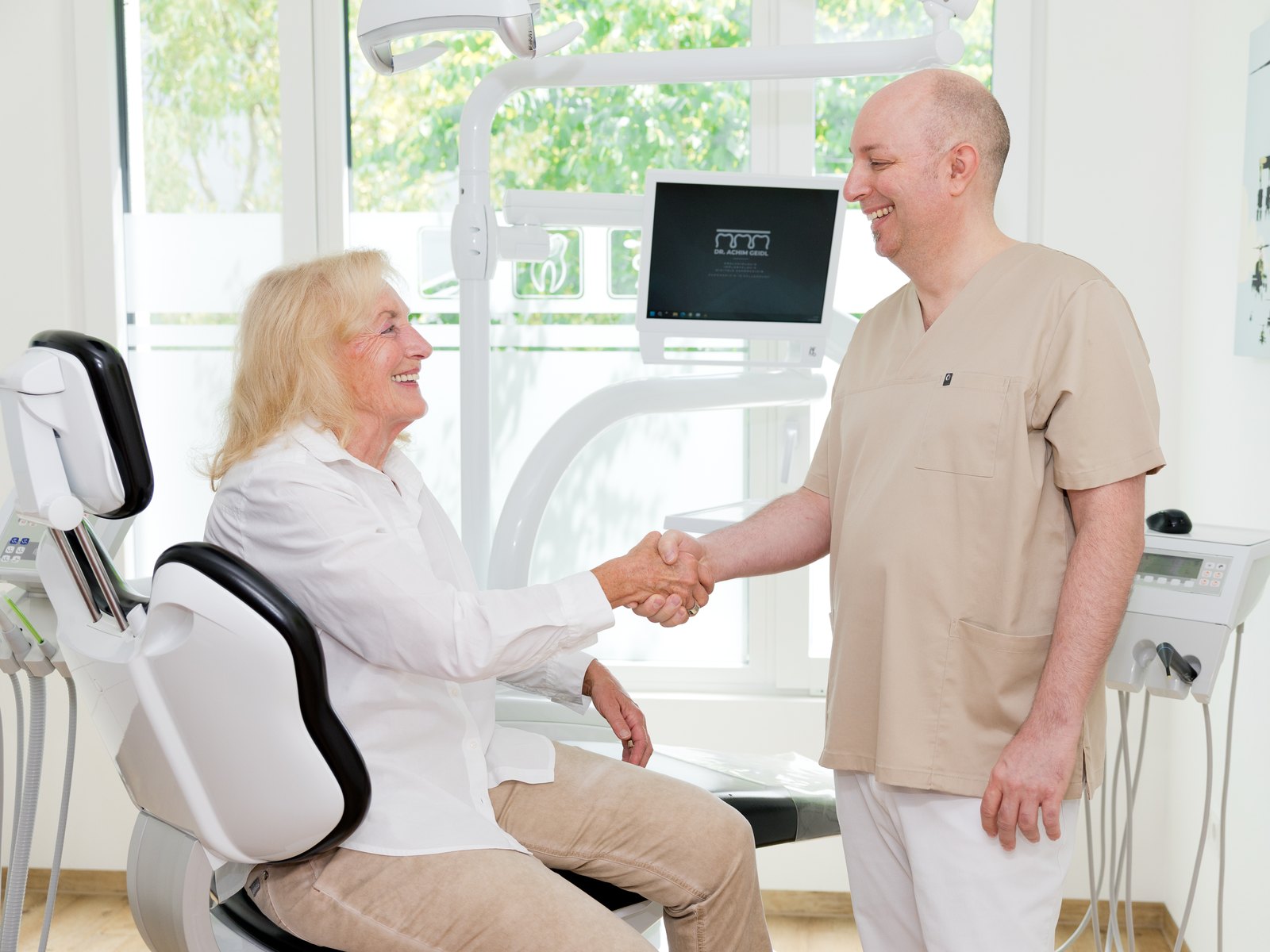 Dr. Achim Geidl begrüßt eine Patientin am Behandlungsstuhl per Handschlag